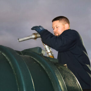 The image shows a man in black suit holding a metal hose beside a green drum.