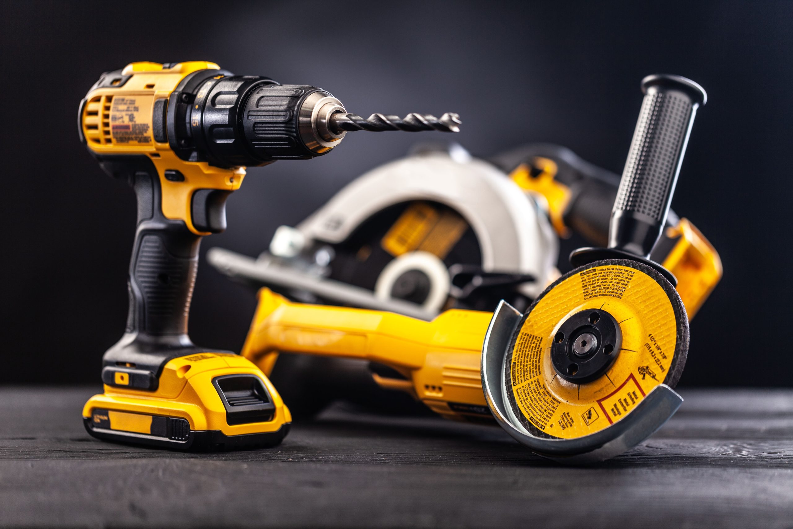 Three yellow-and-black power tools—a cordless drill with bit, circular saw, and angle grinder—arranged on a dark surface, with the drill in sharp focus and the other tools slightly blurred in the background.