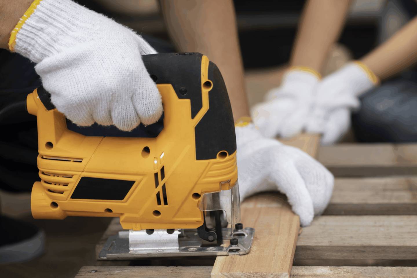 Close-up of a woodworking task where one person uses a yellow and black electric jigsaw to cut a wooden plank placed on a pallet, while another steadies it. Both wear white work gloves, emphasizing safety and precision in a hands-on carpentry project.